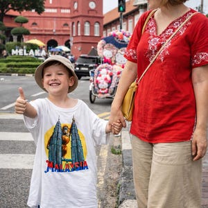 Puede incluir: Un ni&ntilde;o con una camiseta blanca con un gr&aacute;fico de las Torres Petronas en Malasia, pantalones cortos azules y un sombrero beige hace una se&ntilde;al de pulgar hacia arriba. El ni&ntilde;o est&aacute; agarrado de la mano de una persona con una camisa roja con bordados blancos y pantalones beige.