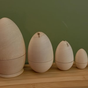 May include: A set of five wooden egg-shaped nesting dolls in a row, each a different size. The dolls are made of natural wood and have a dark line around the base. They are displayed on a light wooden surface, with a green backdrop.