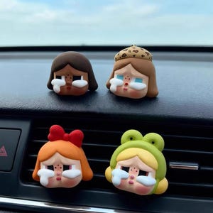 May include: Four colorful car vent clip decorations shaped like cartoon faces with tears. The faces have different hairstyles and accessories, including a red bow, a leopard print hat, and a green frog hat. The decorations are on a black dashboard.