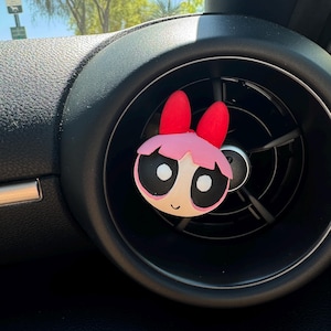 May include: A car air freshener clip featuring the character Blossom from The Powerpuff Girls. The figure has a round face, large black eyes, pink hair, and a red bow on top. It is attached to a circular black plastic car air vent.