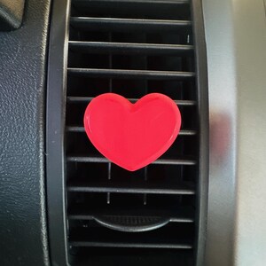 May include: A bright red heart-shaped car air vent clip. The clip is positioned on a black car vent, with a silver-colored dashboard visible in the background. The heart is a solid, glossy color.