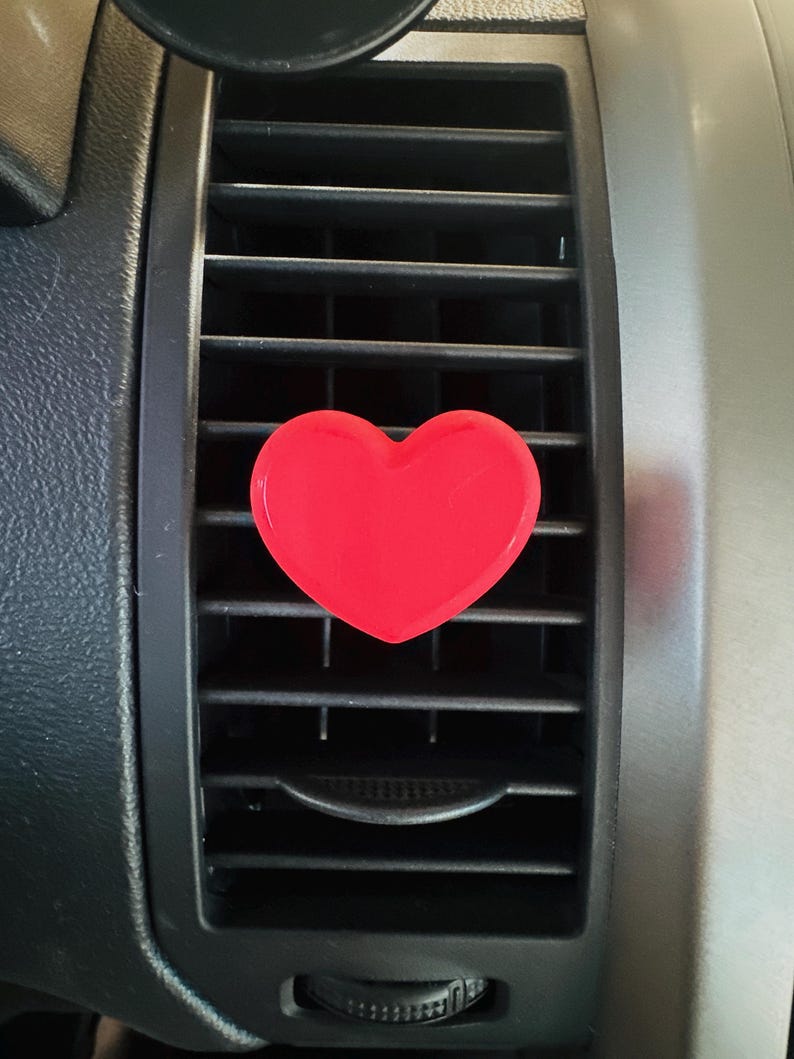 May include: A bright red heart-shaped car air vent clip. The clip is positioned on a black car vent, with a silver-colored dashboard visible in the background. The heart is a solid, glossy color.