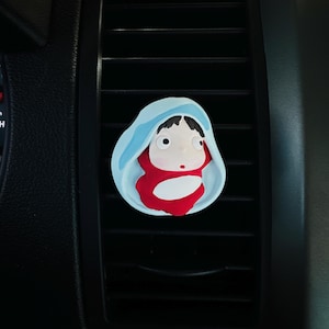 May include: A car air vent clip featuring a cartoon character in a blue hooded cloak and red outfit. The clip is designed to attach to car vents and has a whimsical, artistic design.