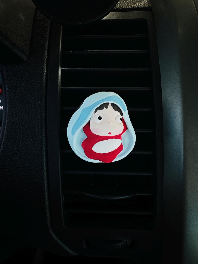 May include: A car air vent clip featuring a cartoon character in a blue hooded cloak and red outfit. The clip is designed to attach to car vents and has a whimsical, artistic design.