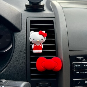 May include: White Hello Kitty car air freshener with a red bow and dress, and a red bow-shaped air freshener. The air fresheners are attached to a car vent. A black phone mount is visible above the vent.