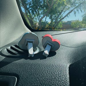 May include: Two car air fresheners. One is shaped like a blue cloud, the other is a red heart. Both have a white clip for attaching to a car vent. The fresheners are sitting on a black dashboard.