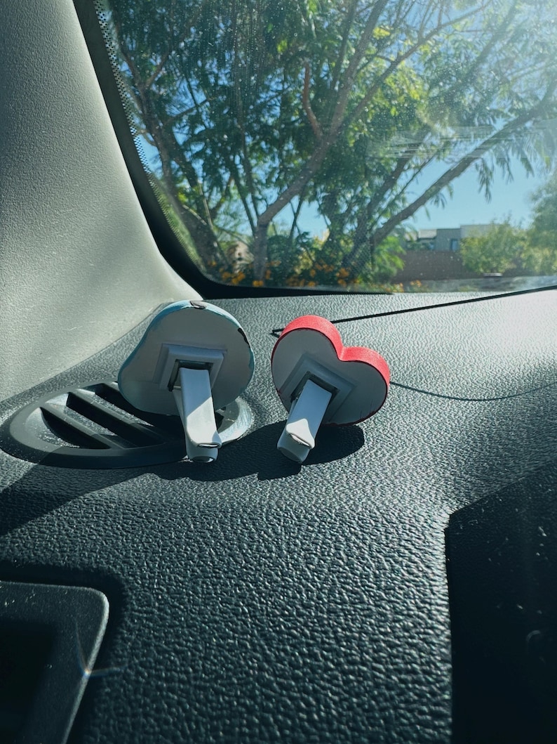 May include: Two car air fresheners. One is shaped like a blue cloud, the other is a red heart. Both have a white clip for attaching to a car vent. The fresheners are sitting on a black dashboard.