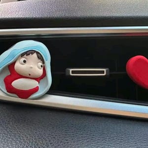 May include: A car air vent decoration featuring a cartoon character in a blue and red outfit, and a red heart. The character is in a light blue frame. The heart is a solid red color.
