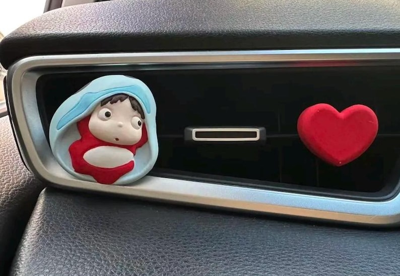 May include: A car air vent decoration featuring a cartoon character in a blue and red outfit, and a red heart. The character is in a light blue frame. The heart is a solid red color.