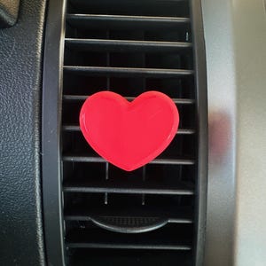 May include: A bright red heart-shaped car air vent clip. The clip is attached to a black car vent with a silver-colored trim. The heart is centered on the vent, providing a pop of color.