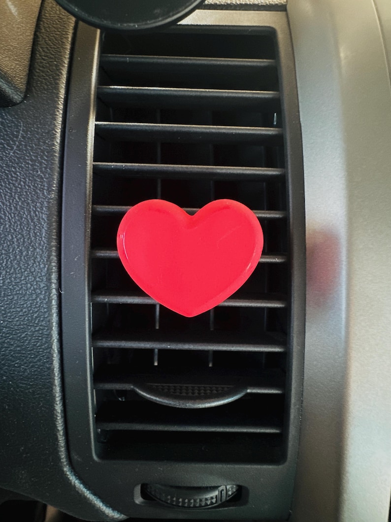 May include: A bright red heart-shaped car air vent clip. The clip is attached to a black car vent with a silver-colored trim. The heart is centered on the vent, providing a pop of color.