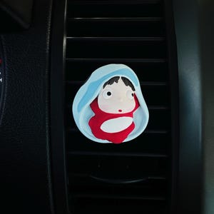 May include: A car air vent clip featuring a cartoon character in a red outfit and a blue hood. The clip is designed to attach to the car's air vent, adding a decorative touch. The character has large eyes and a small red dot on its face.