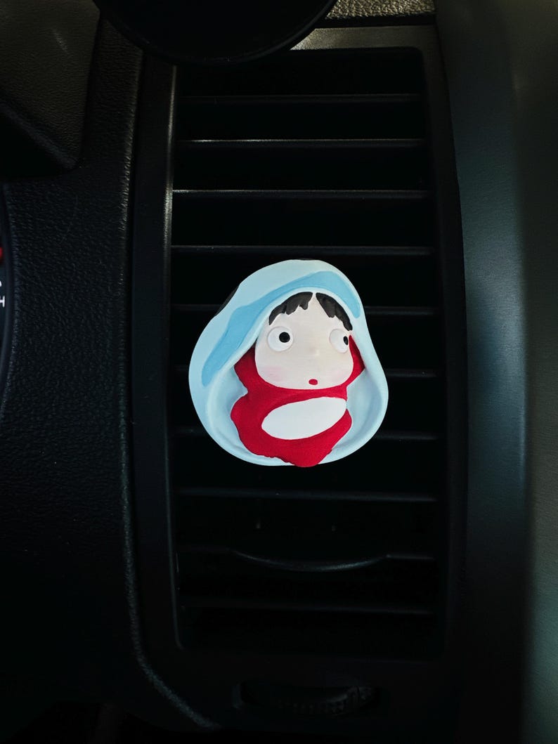 May include: A car air vent clip featuring a cartoon character in a red outfit and a blue hood. The clip is designed to attach to the car's air vent, adding a decorative touch. The character has large eyes and a small red dot on its face.