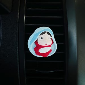 May include: A car air vent clip featuring a cartoon character design. The clip has a light blue hood, a red body, and a white face. The clip is attached to a black car vent.