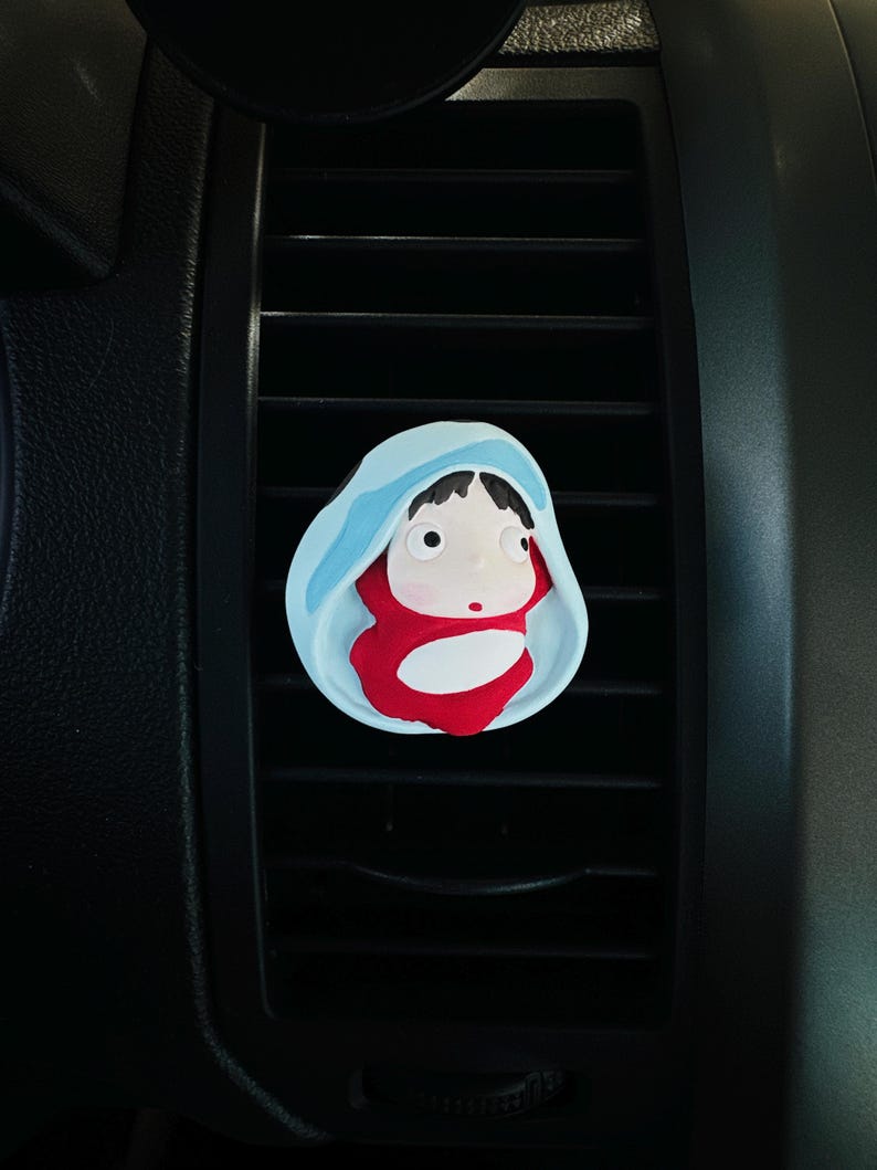 May include: A car air vent clip featuring a cartoon character design. The clip has a light blue hood, a red body, and a white face. The clip is attached to a black car vent.