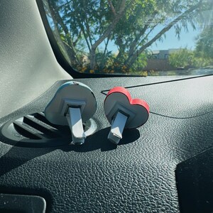 May include: Two car air fresheners. One is shaped like a blue cloud, the other is a red heart. Both have white and gray accents and a clip for attaching to a car vent. The fresheners are on a black dashboard.