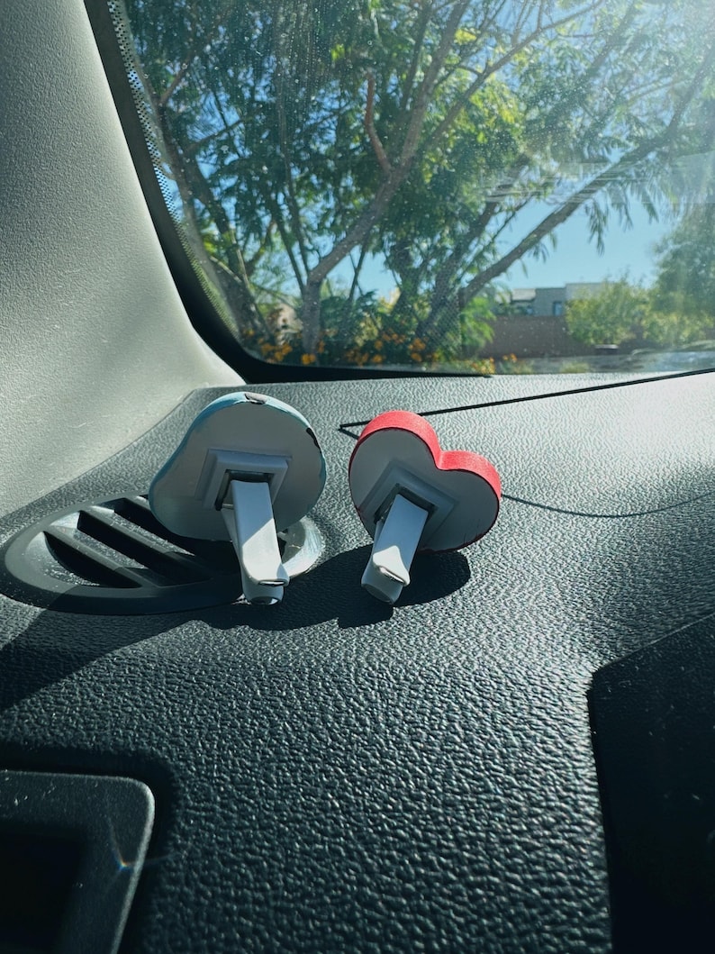 May include: Two car air fresheners. One is shaped like a blue cloud, the other is a red heart. Both have white and gray accents and a clip for attaching to a car vent. The fresheners are on a black dashboard.