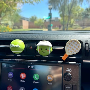 May include: Three car air freshener vent clips featuring a green tennis ball with the word Tennis, a green tennis ball character wearing a white visor, and a cream-colored tennis racket with an orange handle, all attached to a black car dashboard.