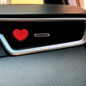 May include: A red heart-shaped car vent clip. The clip is placed on a black car vent with a silver trim. The dashboard is black and textured.