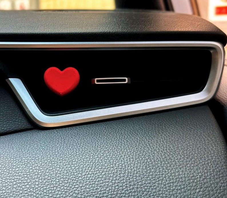 May include: A red heart-shaped car vent clip. The clip is placed on a black car vent with a silver trim. The dashboard is black and textured.