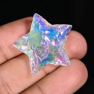 May include: A star-shaped gemstone with an iridescent sheen, displaying colours of blue, green, and purple. The rough-textured stone is held in a hand against a dark backdrop. This unique gemstone could be used as a decorative item or as part of a jewellery piece.