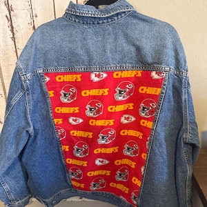 Puede incluir: Una chaqueta de mezclilla azul con un panel rectangular grande en la parte posterior hecho de tela roja. La tela tiene un patr&oacute;n repetitivo de texto amarillo "CHIEFS", logotipos de punta de flecha blancos y rojos e ilustraciones de cascos.