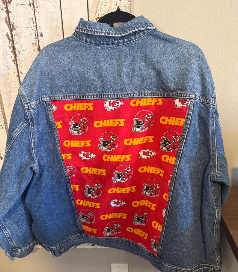 Puede incluir: Una chaqueta de mezclilla azul con un panel rectangular grande en la parte posterior hecho de tela roja. La tela tiene un patr&oacute;n repetitivo de texto amarillo "CHIEFS", logotipos de punta de flecha blancos y rojos e ilustraciones de cascos.
