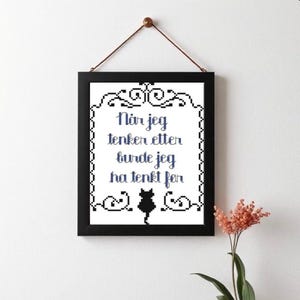 Puede incluir: Arte de pared enmarcado en negro con una cita en texto pixelado azul y negro. La cita está en noruego. Una silueta de gato negro está en la parte inferior. El arte está colgado en una pared blanca con un cordón de cuero marrón.