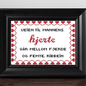 May include: Framed cross-stitch art in a black frame. The white background features red text and a red heart border. The text reads "Veien til mannens hjerte går mellom fjerde og femte ribbein."