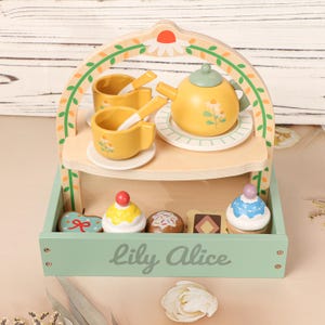 May include: A wooden toy tea set with a yellow teapot, cups, saucers, and spoons. The set includes a tray with cupcakes and cookies. The name "Lily Alice" is printed on the front of the tray.
