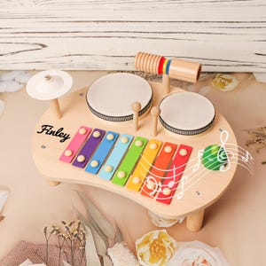 May include: Wooden toy musical instrument set featuring a xylophone with rainbow-colored keys, two drums, a cymbal, and a wooden percussion instrument. The name "Finley" is printed on the instrument. A musical note graphic is also present.