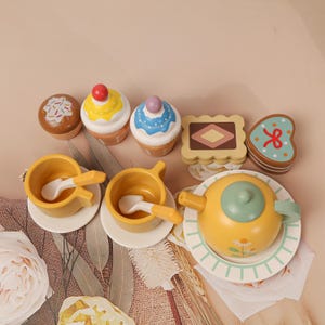 May include: Wooden toy tea set with cupcakes, cookies, teacups, spoons, and a teapot. The cupcakes have yellow, blue, and white frosting with a cherry on top. The teapot is yellow with a green lid and a floral design.