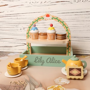 May include: Wooden toy tea set with a two-tiered display stand holding cupcakes and a small cake. The set includes two yellow teacups, a teapot, and a serving tray. The text "Lily Alice" is printed on the tray.