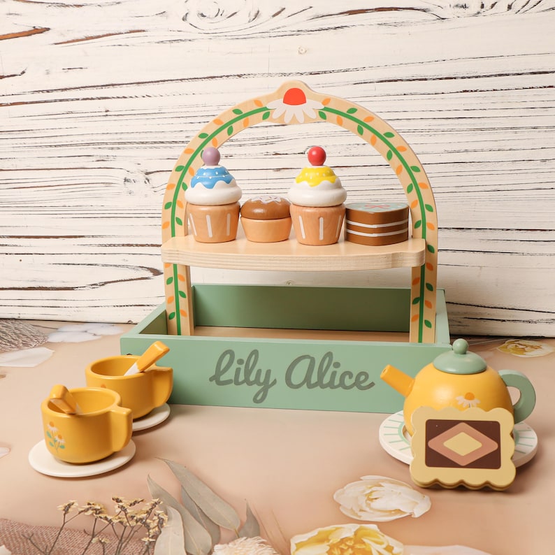 May include: Wooden toy tea set with a two-tiered display stand holding cupcakes and a small cake. The set includes two yellow teacups, a teapot, and a serving tray. The text "Lily Alice" is printed on the tray.