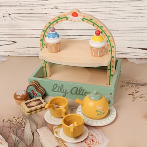May include: A wooden toy tea set with a two-tiered serving tray, cupcakes, and various treats. The set includes a yellow teapot, cups, saucers, and a serving tray with the text "Lily Alice". The tray is light green with a wooden arch.
