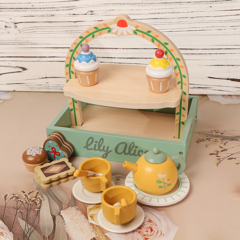 May include: A wooden toy tea set with a two-tiered serving tray, cupcakes, and various treats. The set includes a yellow teapot, cups, saucers, and a serving tray with the text "Lily Alice". The tray is light green with a wooden arch.