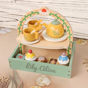 May include: A wooden toy tea set with a yellow teapot, cups, and saucers, along with a tray of toy desserts. The set is painted in pastel colors and includes the text "Lily Alice" on the tray.