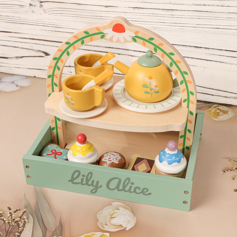 May include: A wooden toy tea set with a yellow teapot, cups, and saucers, along with a tray of toy desserts. The set is painted in pastel colors and includes the text "Lily Alice" on the tray.