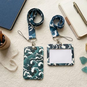 Puede incluir: Dos cordones a juego con porta-identificadores. Los cordones y los porta-identificadores presentan un patrón repetido de olas blancas sobre un fondo verde azulado oscuro. Los porta-identificadores tienen una ventana transparente para mostrar la identificación. Los cordones están conectados a los porta-identificadores con un cierre plateado.