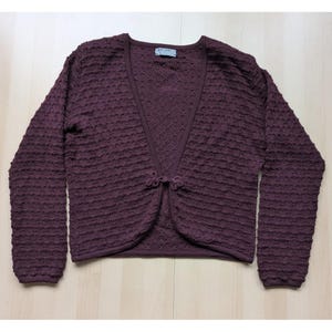 May include: A plum-colored cardigan sweater with a textured, bubble-like knit pattern. The long-sleeved sweater has a V-neck and a decorative closure at the waist. The garment is made of a soft, comfortable material.