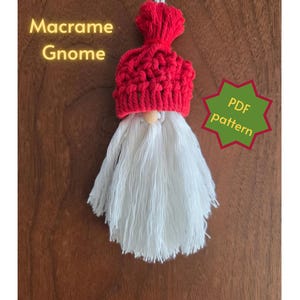 Puede incluir: Un adorno de gnomo de macramé con un gorro rojo anudado con un pompón y una larga barba de flecos blancos. Una pequeña cuenta de madera sirve de nariz. El texto Macrame Gnome y PDF pattern es visible, indicando una guía de patrón digital.