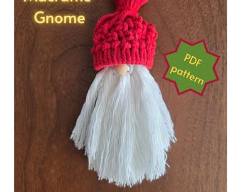 Puede incluir: Un adorno de gnomo de macramé con un gorro rojo anudado con un pompón y una larga barba de flecos blancos. Una pequeña cuenta de madera sirve de nariz. El texto Macrame Gnome y PDF pattern es visible, indicando una guía de patrón digital.