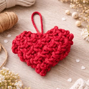 May include: A handmade, red, crocheted heart ornament with a loop for hanging. The heart is made of thick yarn and has a textured appearance. Other items on the wooden surface include white and brown twine, and small white heart-shaped confetti.