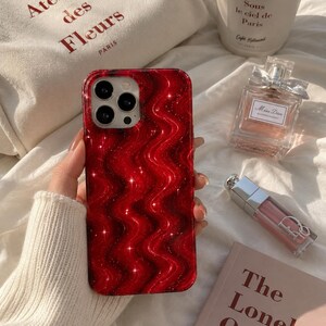 May include: A red phone case with a wavy, glittery design and a camera module. The case is held by a person wearing a white sweater. Other items in the image include a tote bag with text, a coffee cup, perfume, and a book titled "The Lonely Ones."