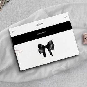 Puede incluir: Una tarjeta rectangular blanca con una franja horizontal negra, una ilustración de lazo en acuarela negra y el texto "SEPTEMBER", "12TH", "2027" y "CLAIRE & MARTIN". Un pequeño clip dorado está sujeto al lateral de la tarjeta.