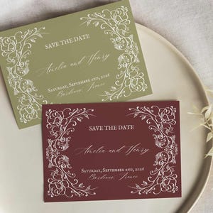 Puede incluir: Dos tarjetas rectangulares de "Save the Date". Una es verde oliva, la otra burdeos. Ambas tarjetas presentan diseños de bordes florales blancos y texto que dice "Save the Date", nombres y una fecha. Las tarjetas están sobre un plato blanco.