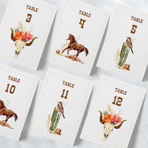 Puede incluir: Un conjunto de tarjetas blancas con números de mesa con un tema rústico y occidental. Cada tarjeta presenta un número marrón, un borde decorativo e ilustraciones de acuarela de un caballo, un pájaro o un arreglo floral con una calavera.