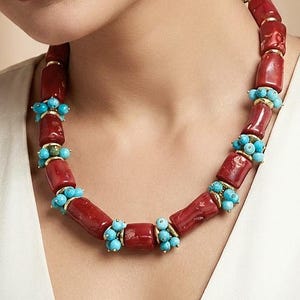 Op de afbeelding: Een opvallende ketting met grote, cilindrische rode koraal kralen afgewisseld met clusters van kleine, ronde turkooizen kralen. De ketting heeft goudkleurige accenten en wordt gepresenteerd tegen een neutrale achtergrond.