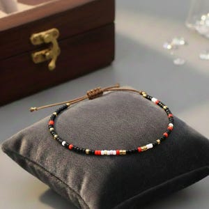 Puede incluir: Una pulsera de cuentas con un dise&ntilde;o de cuentas negras, rojas, blancas y doradas. La pulsera se muestra sobre un coj&iacute;n gris, con una caja de madera marr&oacute;n en el fondo. Tiene un cierre de cord&oacute;n ajustable.