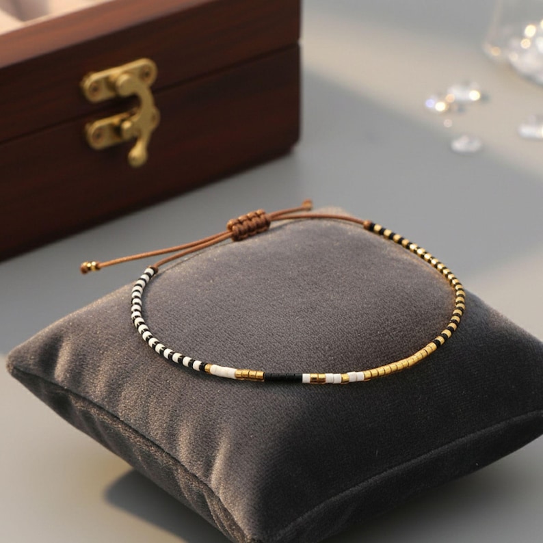 Puede incluir: Una pulsera de cuentas con un cord&oacute;n marr&oacute;n, sobre un coj&iacute;n de terciopelo gris. La pulsera presenta un patr&oacute;n de cuentas negras, blancas y doradas. Una caja de madera con un cierre dorado est&aacute; en el fondo.